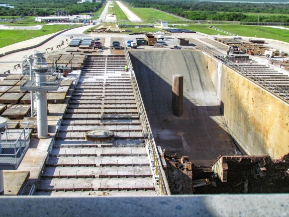 Large-format photograph: Looking south from the North Piping Bridge, the area of Crawlerway Grid Panel refurbishment and replacement can be viewed, along with refurbished and replacement Panels stacked on cribbing adjacent to the work area. In the Flame Trench, work for the Artemis Program is ongoing, and includes the removal of the RSS Rail Beam, which once carried the RSS across the Flame Trench to its mate position on the east side. The column which once supported the Rail Beam remains, and this was the thing that would ”get you” every time, as you attempted to switchback down the too-steep slope on your skateboard after work hours, across the glass-slick surface of the Flame Trench Firebricks. Also, demolition work on the Flame Deflector is visible in the lower right corner of the frame. Photo credit: Withheld by request.